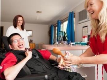 2 women help a man in a wheelchair in a household kitchen.