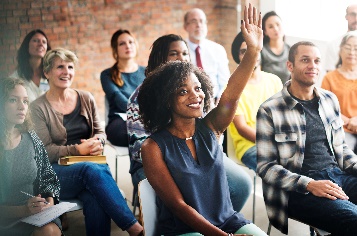 A group of people at a meeting. One person has their hand up to speak. 