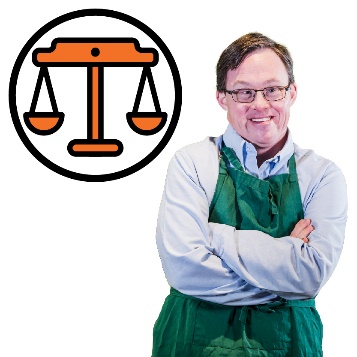 A person wearing green apron and crossing his arms. Next to them is a set of justice scales.
