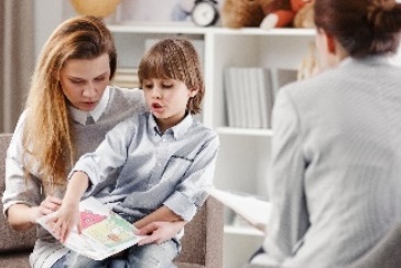 A child with their parent looking through a book together. There is a professional in the room with them. 