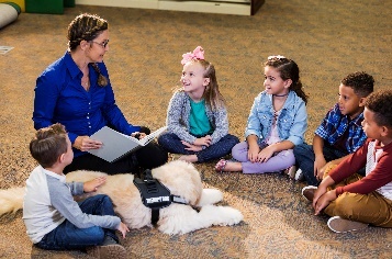 A person talking to a group of children outside on the grass. There is a guide dog. 