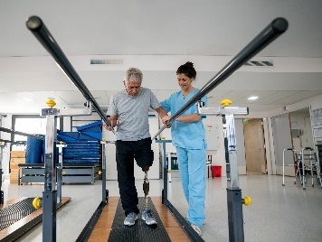 A person with a prosthetic leg being supported by a health care worker to go through physical therapy.