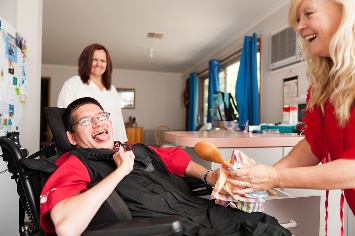 Two women help a man in a wheelchair in a household kitchen.